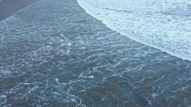 Drone view of a broad ocean wave gliding over dark blue water, creating elegant foam edges, layered textures, and a calm coastal atmosphere.