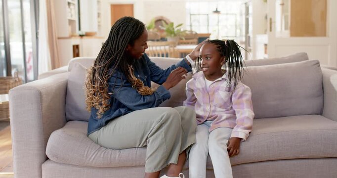 African mother leaning toward child, combing pigtails with comb and ties on light-gray sofa at home