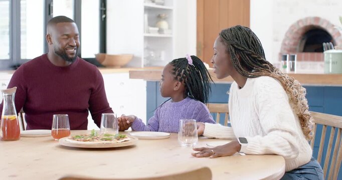 African American family holding hands, father speaking, daughter laughing over shared plate at home