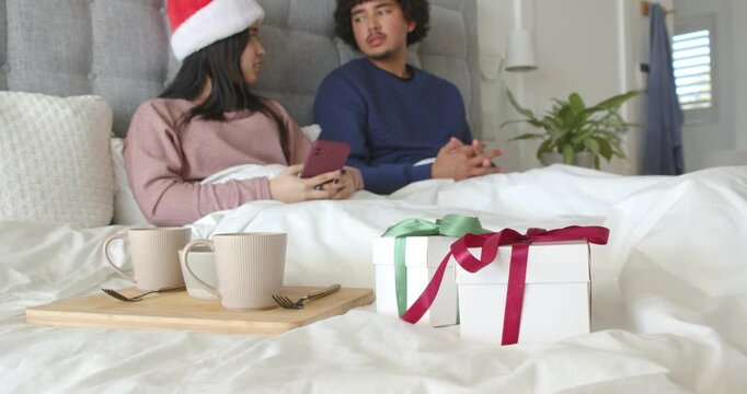 African couple sitting in bed, woman in Santa hat tapping smartphone, man leaning