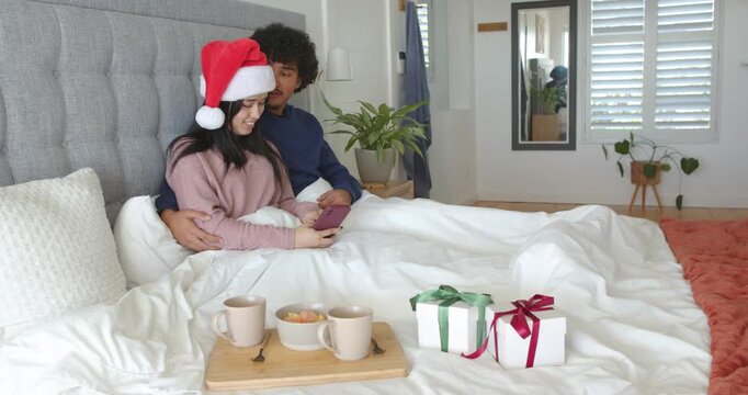 Couple scrolling smartphone on bed during christmas morning, woman in Santa hat, tray gifts visible