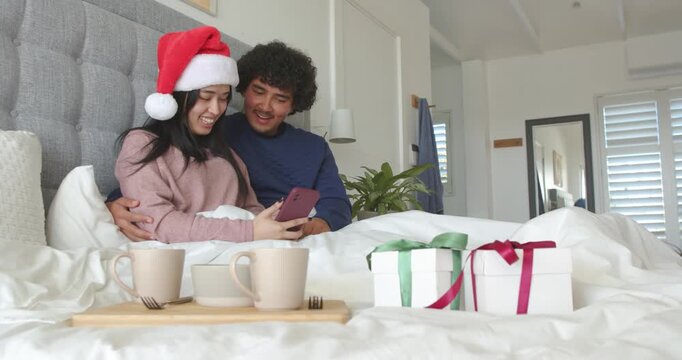 Couple watching christmas message on smartphone on bed, woman wearing Santa hat, viewing gifts