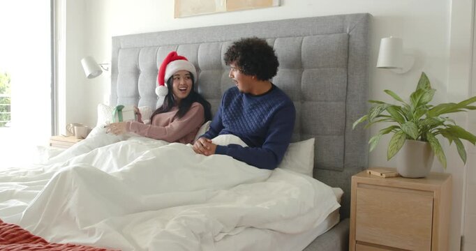 Diverse couple sitting in bed sharing christmas, woman wearing Santa hat offering gift and hugging