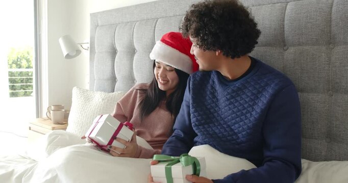 African American couple sitting on bed trading gifts, woman wearing Santa hat celebrating christmas