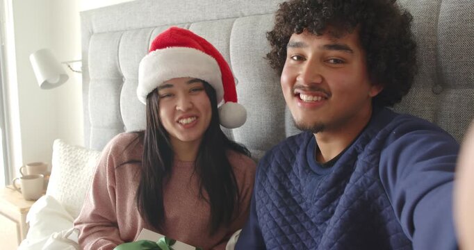 Diverse couple raising camera, filming christmas greeting with woman wearing Santa hat holding gift