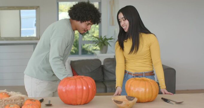 Diverse couple standing at home, comparing pumpkin sizes and arranging carving tools for halloween