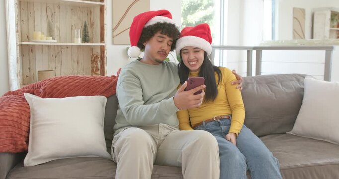 Diverse couple sitting on gray sofa in Santa hats, man shifting smartphone showing christmas clip