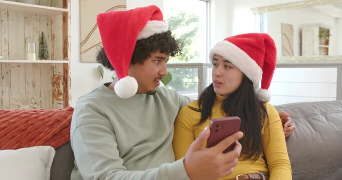 Diverse couple in Santa hats holding smartphone, reacting to christmas call, waving laughing couch