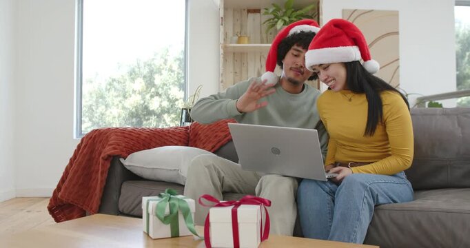 African-American couple in Santa hats taking christmas call, waving, typing on laptop on sofa