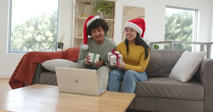 Diverse couple sitting on sofa in Santa hats getting christmas call and showing gifts to laptop