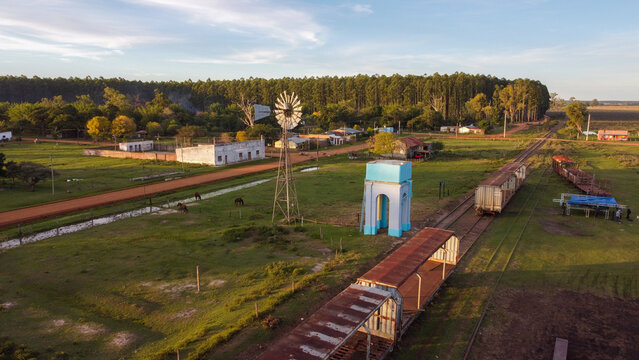 Aerial view of the rural town of Guaviravi featuring a railway line with old train cars, a windmill, and a water tower set amidst green fields in Guaviravi, Corrientes Province, Argentina.