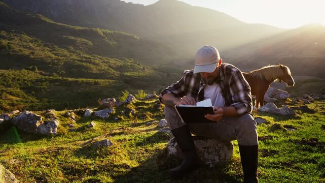 Modern farmer sitting on rock in mountain pasture, reviewing notes on clipboard with horse nearby, combining traditional rural life with professional agricultural work in natural landscape