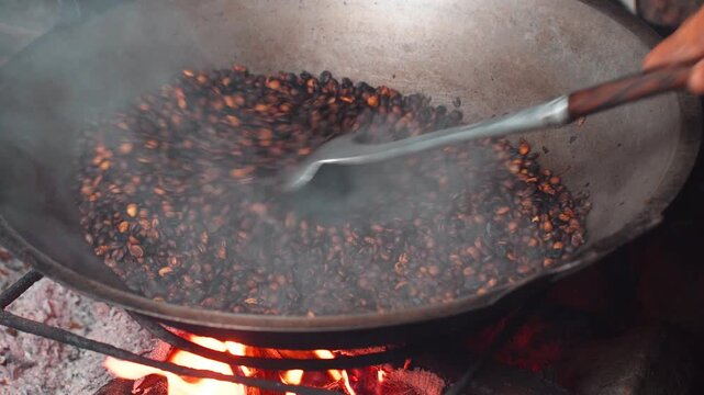 Close up shot of roasting coffee beans in large pan over traditional open wood fire with smoke.
