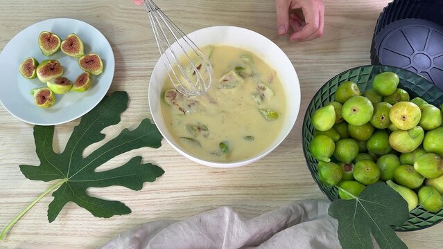 A person uses a metal whisk to mix sliced green figs into a creamy yellow batter inside a white bowl on a wooden table, with whole figs in a green bowl nearby