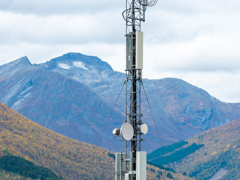 Aerial view of a telecommunications tower with various antennas and equipment, set against a backdrop of rugged mountains and autumn-colored forests in Fjord, More og Romsdal, Norway.