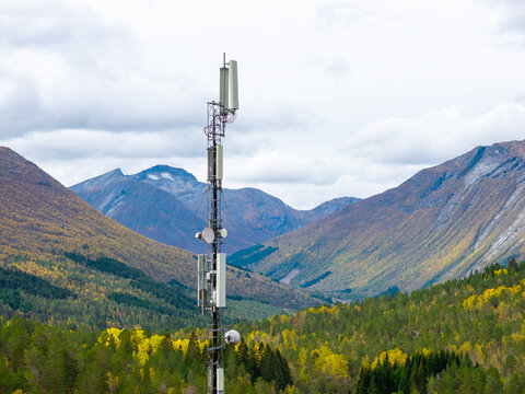Aerial view of a telecommunications tower standing amidst a vast landscape of dense pine forests and towering mountains under a cloudy sky in Fjord, More og Romsdal, Norway.