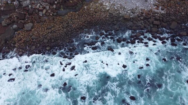 Aerial shot of foamy turquoise waves flowing over a dark rocky coast, combining textured water, scattered stones, and natural coastal movement.