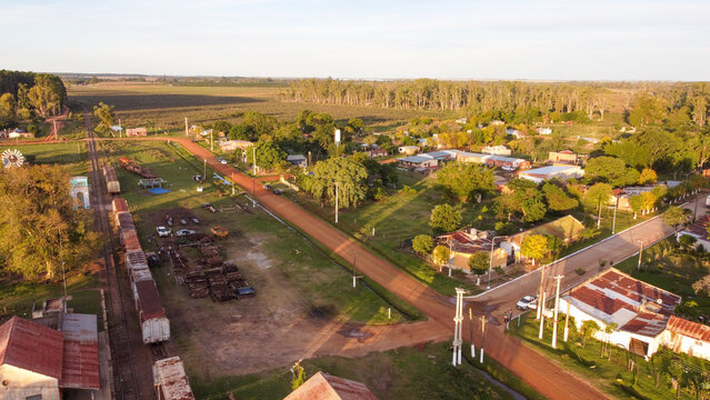 Aerial view of the rural town with a railway line, freight cars, dirt roads, houses, and agricultural fields under warm golden hour light in Guaviravi, Corrientes Province, Argentina.