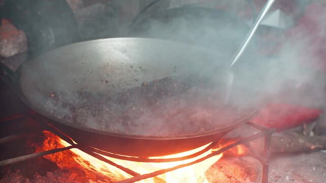 Close up shot of roasting coffee beans in large pan over traditional open wood fire with smoke.
