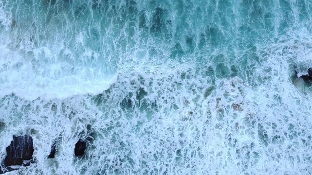 Close aerial view of a powerful turquoise wave exploding into white foam, revealing dark rocks and energetic ocean movement.