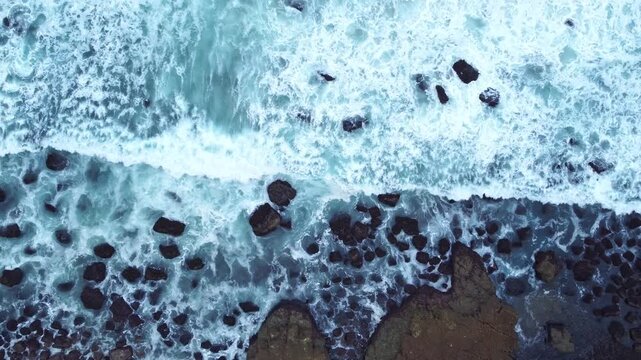 Overhead coastal footage showing white surf swirling around dark rocks, with clear blue water and intricate natural shoreline patterns.