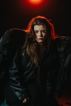A young woman posed wearing black wings and a leather jacket in dramatic lighting. Moody studio portrait with red backlight highlighting her hair and dark costume.
