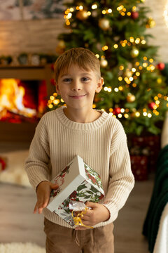 Cute preschool boy, sitting in front of the fireplace, holding christmas present and reading a book, joy and happiness