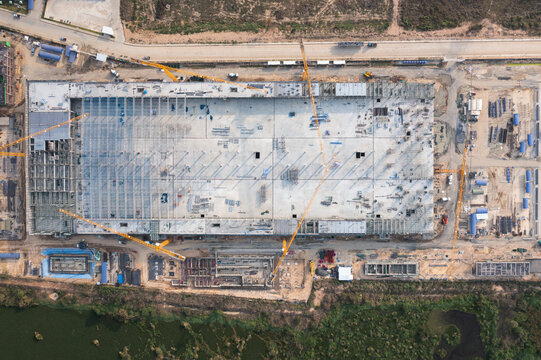 Top-down drone view of a large industrial construction project with a broad concrete floor deck, tower cranes, steel grid framework, site workers, equipment and material staging beside an access road.