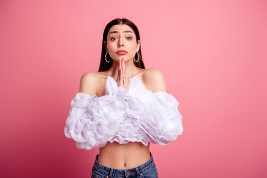 Young woman pleading with hands together in prayerful pose wearing ruffled white blouse against pink background expressing hope and apology