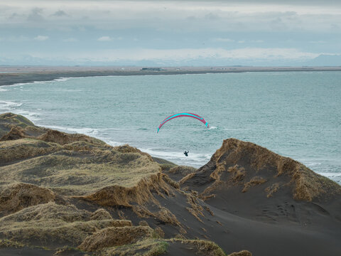Aerial view of a paraglider soaring over the black sand dunes of Hafnarskeidh with the ocean and distant mountains in the background in Sveitarfelagid Olfus, Iceland.