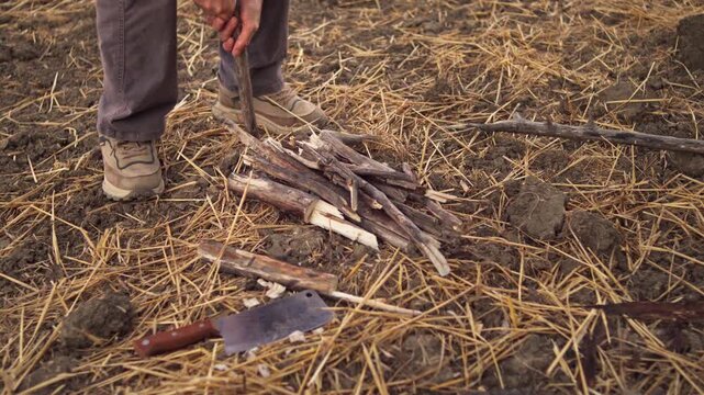 A person pushes a sharpened wooden pole into the ground beside a pile of dry kindling sticks and a cleaver knife in a field, medium shot.