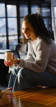 Vertical video: African American woman chatting with pal holding mug at cafe table in gray top