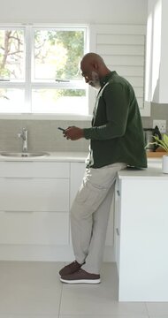 Vertical video: Mature African man leaning on counter in kitchen, tapping phone and checking texts