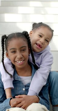 Vertical video: Two African female children sitting on steps smaller climbing behind hugging older