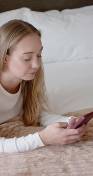 Vertical video: Woman propping on forearms, scrolling pink-cased smartphone on bed, copy space