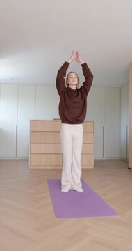 Vertical video: Female standing on purple mat at home inhaling, raising arms into prayer for calm