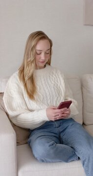 Vertical video: Woman in sweater sitting on sofa with pink phone scrolling then placing on sofa arm