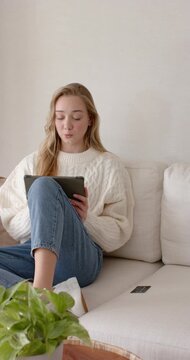 Vertical video: Female in cream sweater, jeans sitting on sofa, reading tablet, reacting to content