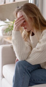 Vertical video: Adult woman in jumper leaning on sofa pressing head then calming gazing at window