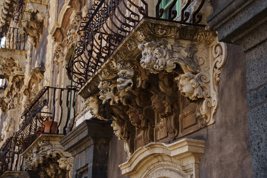 Ornate baroque balcony in Sicily with elaborate stone carvings of faces, cherubs, and aged masonry textures with wrought iron railings at San Nicolo l'Arena Benedictine Monastery in Catania