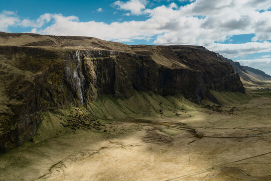Aerial view of Seljalandsfoss waterfall cascading down a dramatic cliff face in a vast, grassy landscape under a bright blue sky with scattered clouds in Rangarthing eystra, Iceland.