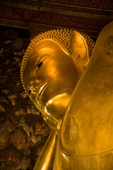 Close-up of the serene golden face of the famous giant Reclining Buddha statue with ancient mural background in Wat Pho temple, Bangkok, Thailand. © VaZu Photo