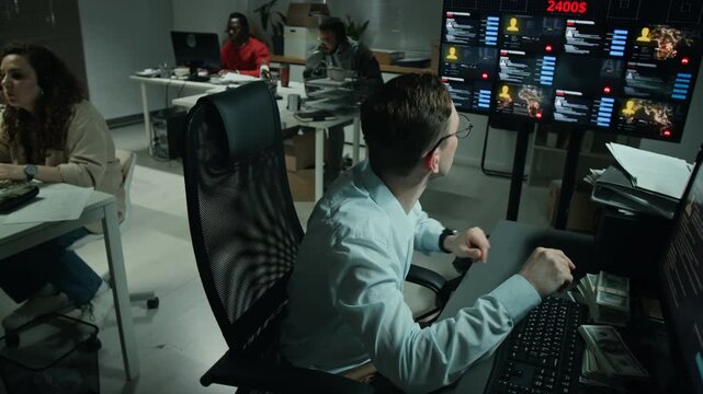 Young Caucasian man monitoring financial data screens, celebrating successful online transaction with fist pump in scam office. Footage suits cybersecurity videos
