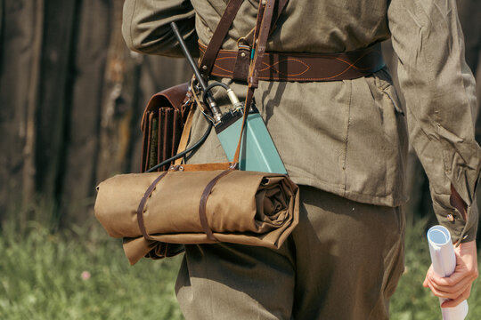 A person wearing a vintage military uniform carries a portable radio transceiver, a brown leather map case, and a rolled canvas mat
