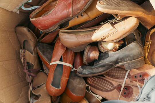 A top-down view shows a high volume of old worn vintage leather shoes and sandals packed randomly inside a cardboard storage box.