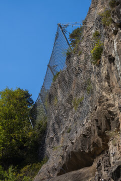 Steel wire mesh rockfall barrier installed on a steep rocky cliff face for slope stabilization and geological hazard prevention, with blue sky and green trees