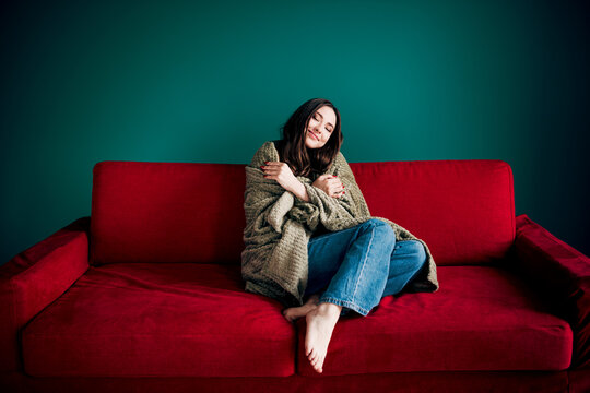 Woman wrapped in blanket relaxing on a red couch at home, cozy comfort and contentment in a casual hygge moment