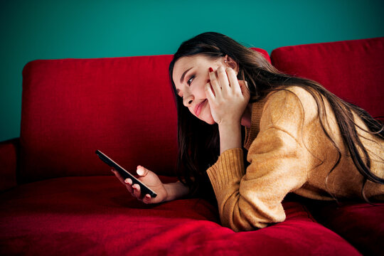Young woman lying on red sofa at home, casually scrolling smartphone and relaxing in a cozy sweater