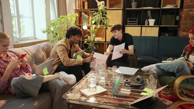Two male colleagues review printed documents while women manage devices at creative workspace with concentrated attention. Document review, task coordination, distributed teamwork concept
