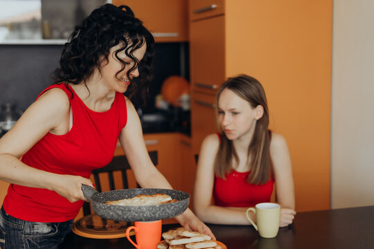 Two women share kitchen space while one serves homemade food and another sits with quiet expression creating domestic scene on emotional labor shared routines household wellbeing and mindful living.